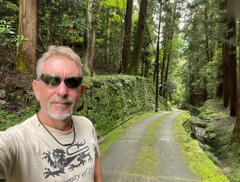 Scott on a green forest path in Japan, wearing sunglasses and a University of Sydney T-shirt